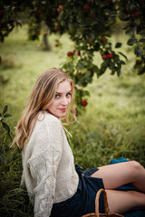 Blonde woman is sitting under a tree full of red apples. She is wearing a cozy sweater and a skirt, enjoying warm autumn evening in a garden