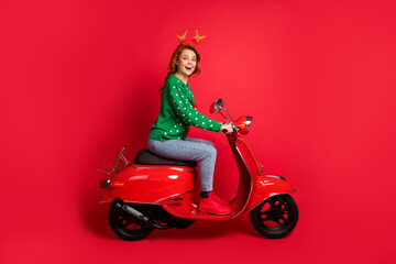 Fototapeta na wymiar Photo portrait side view of woman riding scooter wearing green jumper deer toy headwear isolated on vivid red colored background