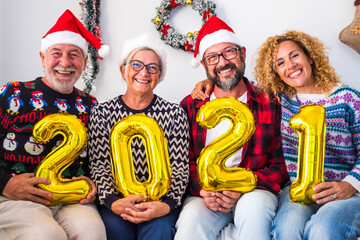 group of four happty and cheerful people holding golden numbers of the new year 2021 at home wearing christmas clothes and hats or caps - four young and mature people having fun celebrating