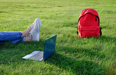 Freelance business concept. Woman legs on green grass lawn in city park, laptop pc computer and backpack. Lifestyle authentic student girl studying outdoors. Mobile office