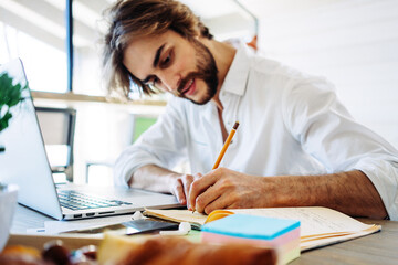 Handsome man with stubble sitting at home and working remote using laptop, making notes with...