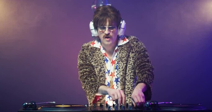 Retro style Caucasian male DJ touching vinyl and dancing while mixing music on stereo equipment. Vintage man in leopard jacket with headphones djing at party at nightclub. 60s disco concept