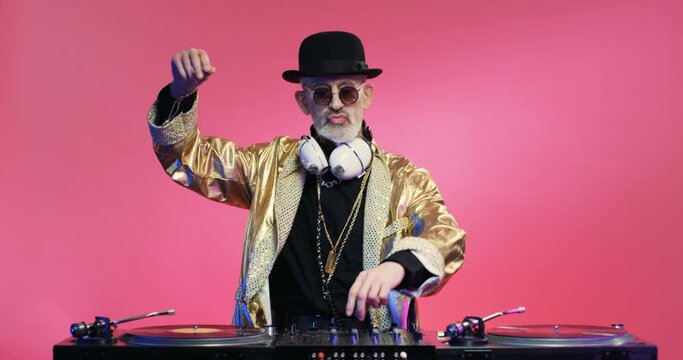 Funny stylish retro Caucasian man in golden sparkling coat and hat moving in rhythm of music and spinning record on musical equipment. Vintage DJ work. Fancy grandpa at mixer on pink wall background.