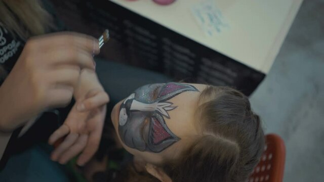 woman draws face painting aqua makeup to little girl