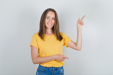 Young woman pointing fingers away in yellow t-shirt, shorts and looking cheerful , front view.