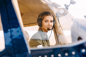 Woman pilot sitting in airplane cockpit, wearing headset, looking at camera, smiling.