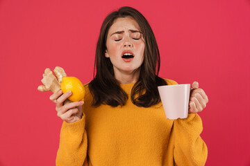 Image of ill caucasian girl holding lemon and ginger while sneezing