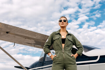 Woman pilot wearing overall and sunglasses, standing next to a private plane on blue sky background.