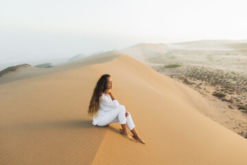 Fototapeta na wymiar Beautiful brunette woman in white clothes relaxing on top of sand dune in Morocco Sahara desert. Mist in morning before sunrise. Harmony with nature.