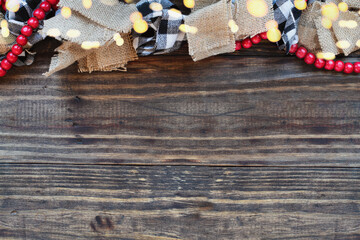 Christmas garland with bokeh lights, burlap, black and white buffalo check fabric and a string of red beads over rust wood table background. Top view with copy space available.