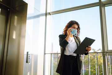 Fototapeta premium Young business woman in protective sterile mask talking on mobile phone while standing near modern elevator. Work during pandemic in quarantine city. Covid-19.