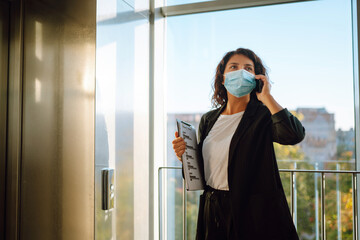 Young business woman  in protective sterile mask talking on mobile phone while standing near modern elevator. Work during pandemic in quarantine city. Covid-19.