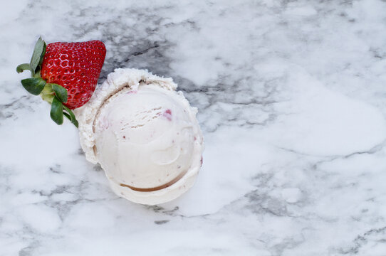 Flatlay view of one scoop of pink strawberry ice cream with a single fresh strawberry fruit over a white and grey marble background texture. Top view.