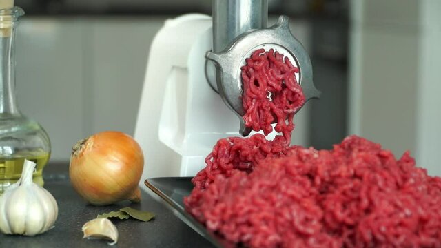 Chef hands put meat in electric meat grinder on background with onion, garlic and olive oil. Preparation of minced fresh beef for burgers. Food background.