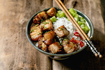 Grilled deep fried pork belly in bowl with rice, celery, chili pepper and spring onion with chopsticks over old wooden background.