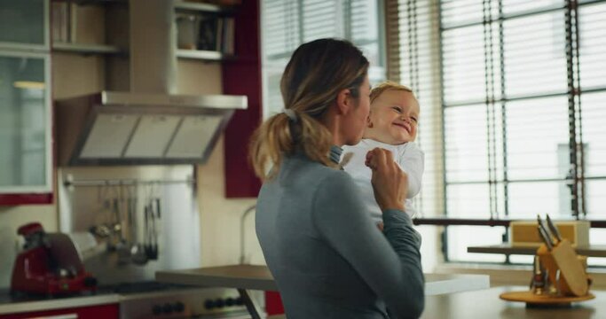 Authentic shot of an young happy smiling mother is keeping in arms her toddler baby boy and having fun to dance together in a kitchen at home.