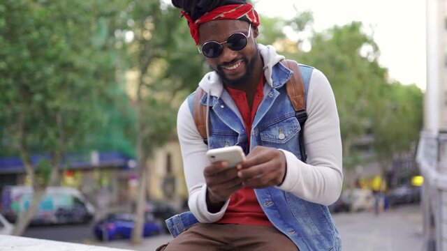 Stock photo of happy african american boy with dreadlocks and a bandana walking in the street. He is using his phone.