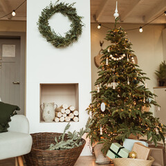 Beautiful Christmas tree, wreath frame, gift boxes in cozy comfortable living room. Festive Christmas / New Year holidays celebration decorations in modern home interior design with sofa and carpet.