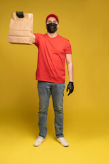 Fototapeta premium Man in red cap, t-shirt in protective mask and gloves giving fast food order isolated on yellow background. Male employee courier hold empty paper packet with food