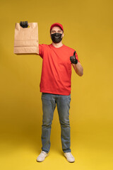 Man in red cap, t-shirt in protective mask and gloves giving fast food order isolated on yellow background. Male employee courier hold empty paper packet with food