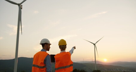 Back view on the man and woman in helmets standing at the electric windmills and discussing something.