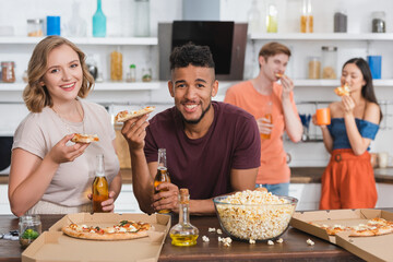 pleased multiethnic friends looking at camera while eating pizza during party
