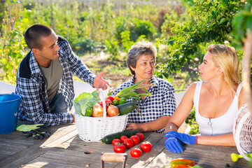 Smiling elderly woman with two adult children breezily chatting at wooden table in garden on summer day happy with rich harvest of vegetables