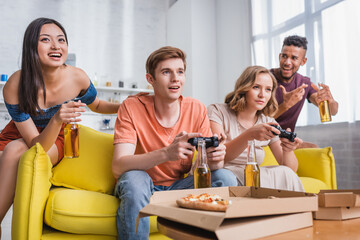 KYIV, UKRAINE - JULY 28, 2020: excited asian woman and african american man holding bottles of beer near friends playing video game