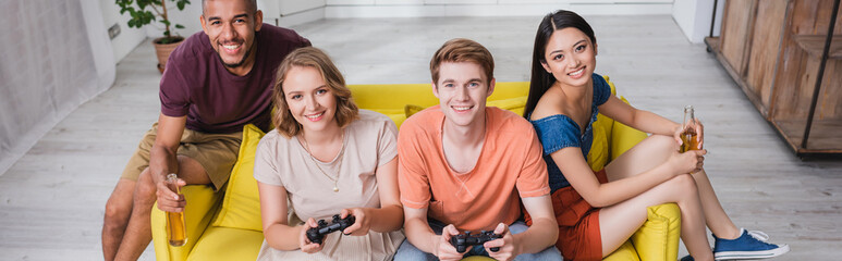 KYIV, UKRAINE - JULY 28, 2020: high angle view of multicultural friends holding beer and playing video game in kitchen, panoramic crop © LIGHTFIELD STUDIOS