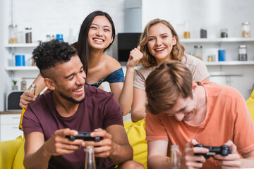 Fototapeta premium KYIV, UKRAINE - JULY 28, 2020: selective focus of excited asian woman showing winner gesture near multicultural friends playing video game