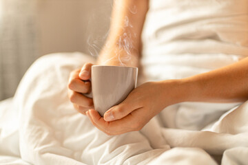 Cup of coffee in hands in bed on a white blanket in the rays of sunlight. Lifestyle concept.