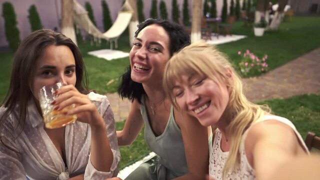 Three joyful women met in veranda cafe outdoors taking selfie photo or video on smartphone, having fun, drink