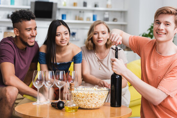 young man opening bottle of red wine near multicultural friends