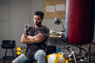In the garage, a car mechanic sits on a yellow motorcycle that he has repaired. Pumped up stylish...