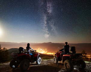 Back view of man and girl trevelers riding on atv quad motorbikes on the top of mountain, romantic couple enjoying beautiful view of night sky full of stars, Milky way, luminous city on background © anatoliy_gleb