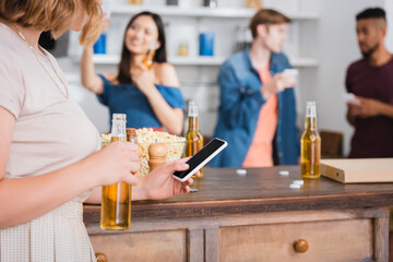 selective focus of young woman holding beer and using smartphone near multicultural friends