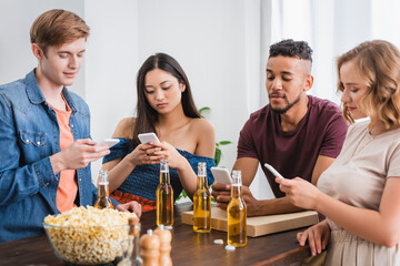 selective focus of multiethnic friends messaging on smartphones near beer and popcorn during party