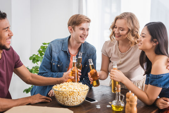 Excited Multicultural Friends Clinking Bottles Of Beer Near Popcorn During Party