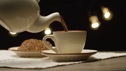 Close up hot tea pouring into a white cup. Blurry lights on black background. Steam rises above the...