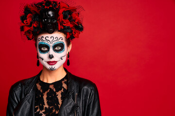 Fototapeta na wymiar Young creepy lady calavera. wears artistic make-up for the feast of all the dead. dressed in a black leather jacket with a wreath of flowers on her head.Symbol of the Day of the Dead isolated in red.