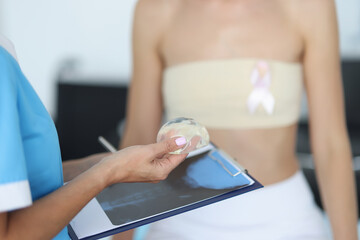 Doctor's hands are holding breast implant in front of clipboard with patient in background....