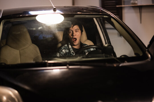 A Young Man Yawns At The Wheel Of An Old Black Car Parked In The Garage