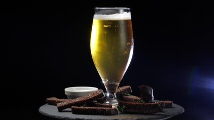 Light chilled cold beer in glass and pieces of dark bread croutons on black wooden background, food composition rotates on circle. Lot of foam and bubbles in mug in alcoholic ale, drops roll down - Powered by Adobe