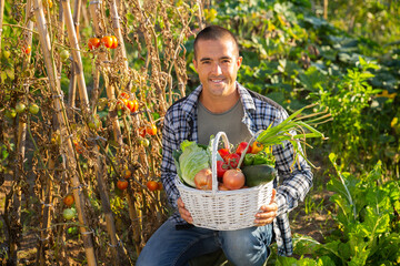 Fototapeta premium Young positive man posing with harvest of vegetables in garden, harvesting season