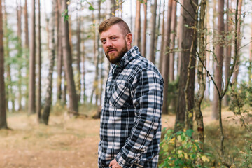 Naklejka na ściany i meble Bearded man in plaid shirt standing in a forest looking away. 