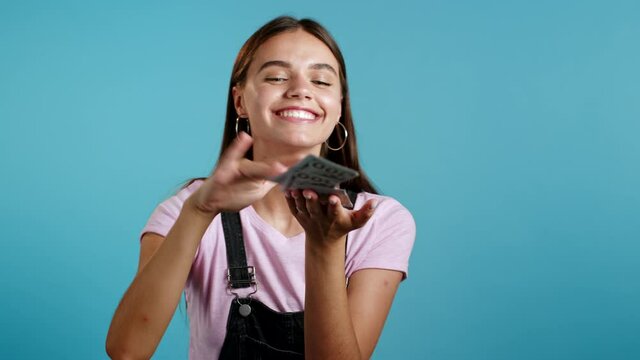 Pretty woman with happy face scatters, waste money. Girl overspend US currency. Lady is flush with dollars on blue studio background .