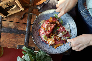 A female hand is preparing a vegetable salad with beef roast beef. A plate of salad with beef roast beef, onions, arugula, pepper and tomatoes on a black table. Top view.