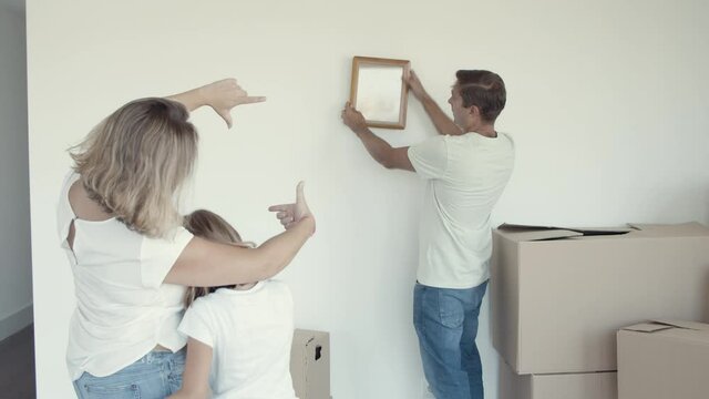 Girl with her mom helping dad to choose place for picture frame on wall. Family establishing new home together. Moving into new apartment concept