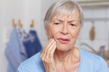Acute toothache: senior woman in her kitchen with a hand at her painful jaw.