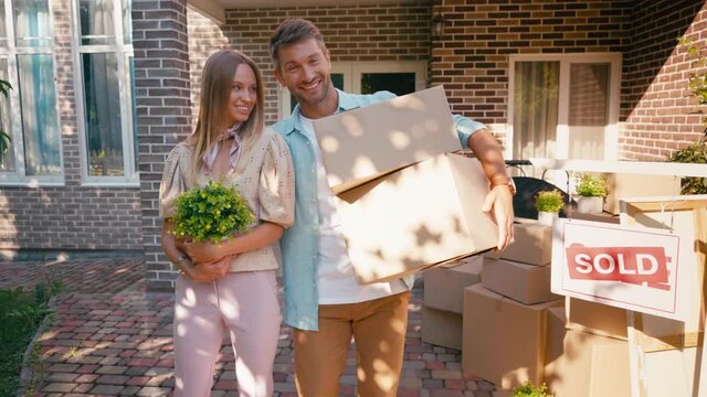 pleased couple holding carton boxes and plant near new house